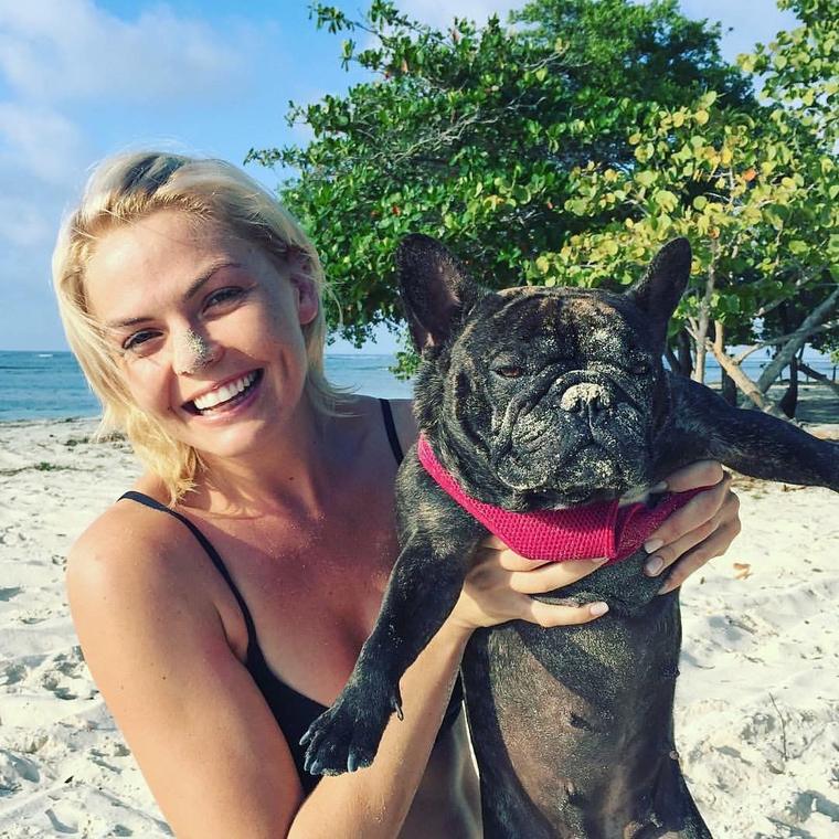 Amy on a white-sand beach in Honduras holding up a sandy brindle French bulldog