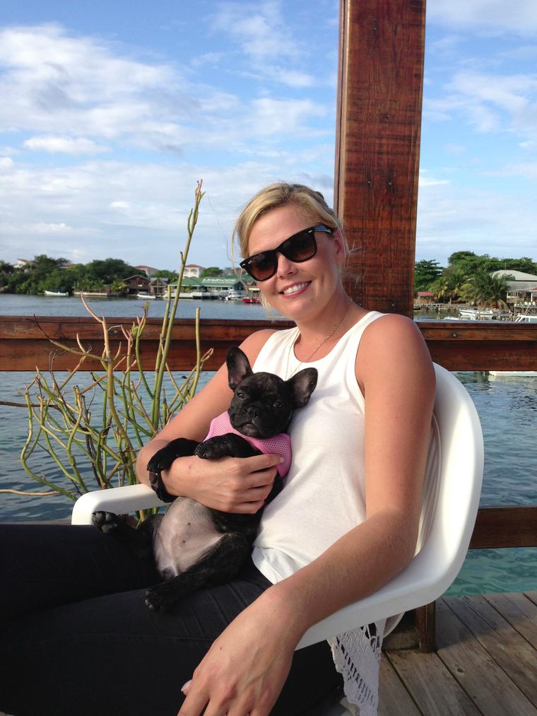 Amy on a waterfront deck cuddling Cosquilla as a brindle French bulldog puppy in a pink harness